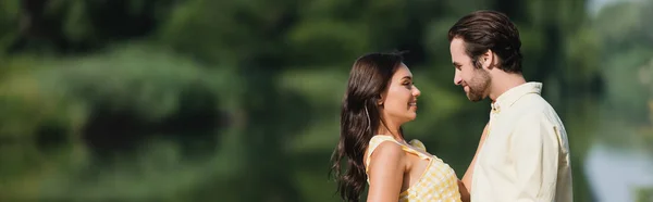 Vue latérale du jeune couple heureux se regardant près du lac, bannière — Photo de stock