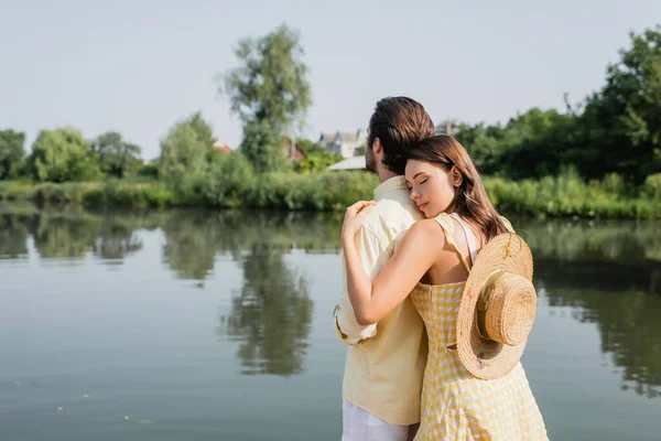 Giovane donna con cappello di paglia abbracciare fidanzato vicino lago — Foto stock
