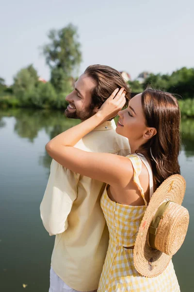 Jeune femme avec chapeau de paille caressant les cheveux de petit ami joyeux près du lac — Photo de stock