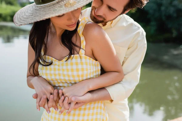 Bearded man hugging and holding hands with girlfriend in straw hat near lake — Stock Photo