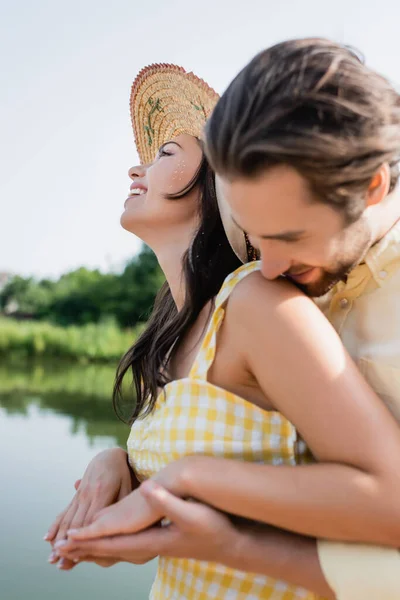 Hombre barbudo besando hombro de novia feliz en sombrero de paja cerca del lago - foto de stock
