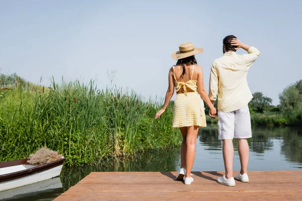 Vista trasera de la joven pareja en ropa de verano cogida de la mano y de pie en el muelle cerca del lago - foto de stock