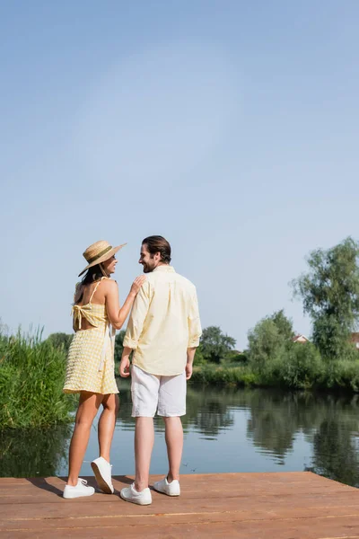 Full length of happy young couple in summer clothes holding hands and standing on pier near lake — Stock Photo