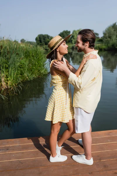 Full length of happy young couple in summer clothes hugging on pier near lake — Stock Photo