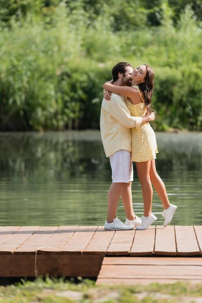 Full length of joyful young couple in summer clothes hugging on pier near lake — Stock Photo