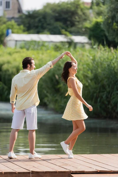Full length of joyful young couple in summer clothes dancing on pier near lake — Stock Photo