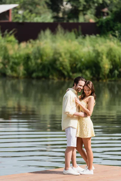 Full length of pleased young couple in summer clothes hugging on pier near lake — Stock Photo