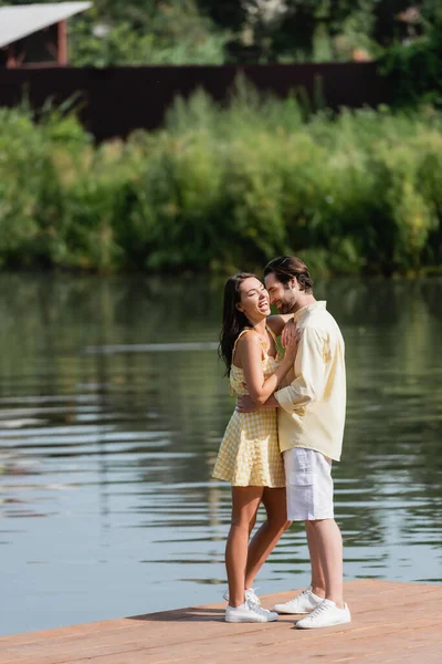 Full length of positive young couple in summer clothes hugging on pier near lake — Stock Photo