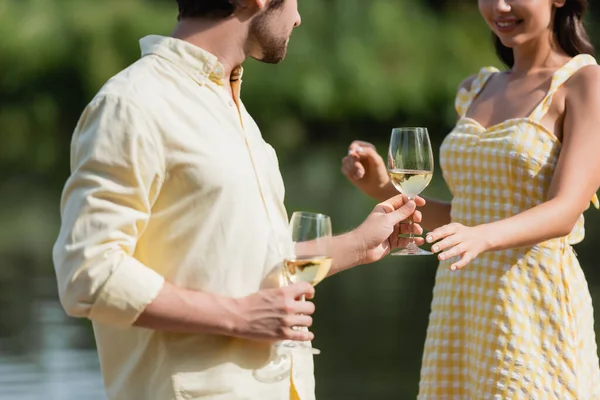 Cropped view of happy young couple in summer clothes holding glasses with wine near lake — Stock Photo