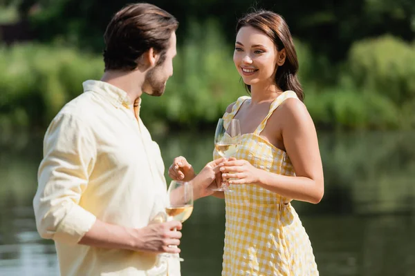 Feliz jovem casal em roupas de verão segurando copos com vinho perto do lago — Fotografia de Stock