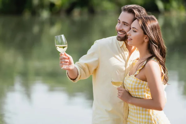 Smiling young couple in summer clothes holding glasses with wine near lake — Stock Photo