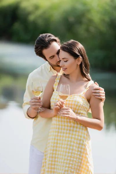 Romántico joven pareja en sonriendo y sosteniendo vasos con vino cerca del lago - foto de stock