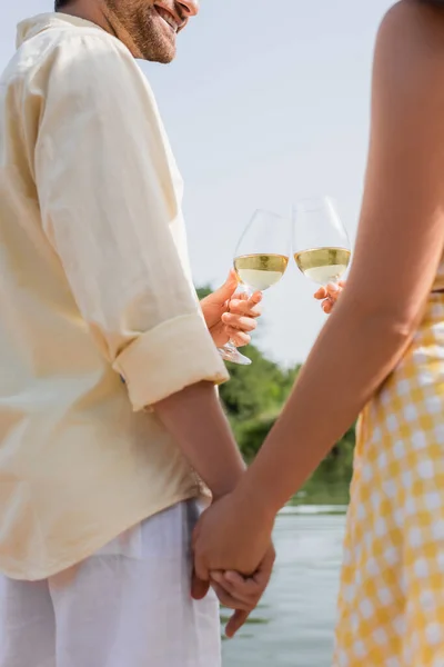 Vista recortada de feliz hombre y mujer tintineo vasos con vino y tomarse de la mano - foto de stock