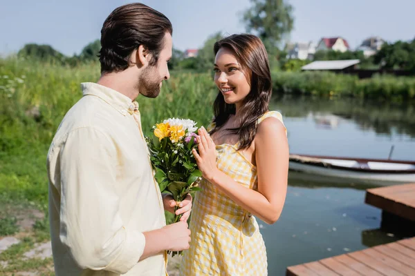 Barbudo hombre celebración ramo de flores cerca de mujer feliz en vestido cerca del lago - foto de stock