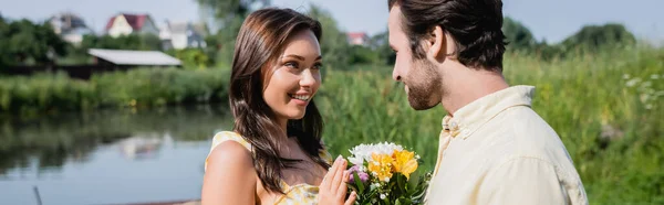 Hombre barbudo sosteniendo ramo de flores cerca de mujer feliz en vestido cerca del lago, bandera - foto de stock