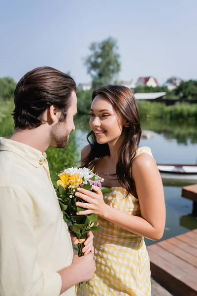 Uomo barbuto tenendo bouquet di fiori vicino donna allegra in abito vicino lago — Foto stock