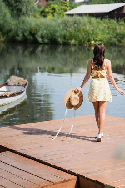 Back view of young woman in summer dress holding straw hat near lake — Stock Photo