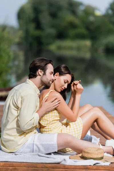 Hombre abrazando sensual joven mujer mientras está sentado en el muelle cerca del lago - foto de stock