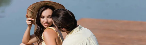 Homme regardant jeune femme souriante en chapeau de paille près du lac, bannière — Photo de stock