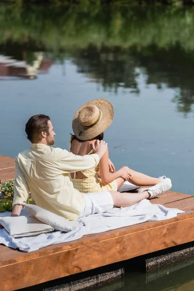 Barbudo hombre y mujer en sombrero de paja sentado en el muelle durante el picnic - foto de stock