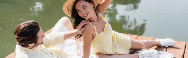 Uomo guardando donna sorridente in cappello di paglia mentre seduto sul molo vicino al fiume, striscione — Foto stock