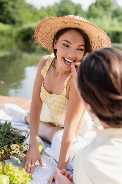 Blurred man feeding smiling woman in straw hat with grape during picnic — Stock Photo