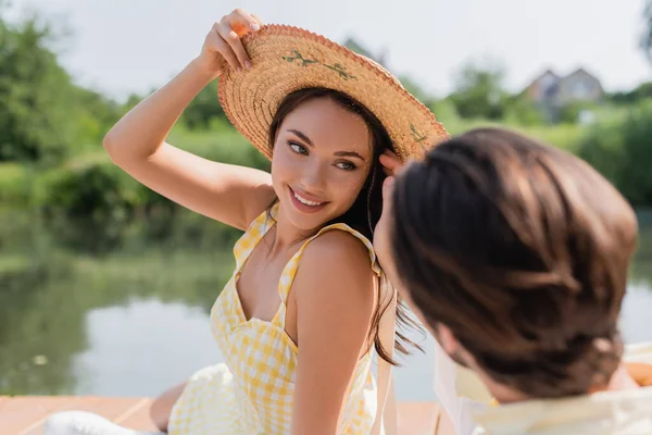 Размытый мужчина смотрит на улыбающуюся женщину, поправляющую соломенную шляпу у реки — стоковое фото
