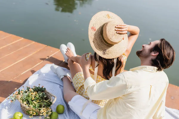 Vista de ángulo alto del hombre alegre mirando a la novia en sombrero de paja mientras está sentado en el muelle durante el picnic - foto de stock