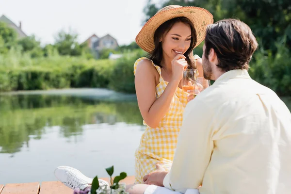 Aufgeregte Frau mit Strohhut hält ein Glas Wein in der Hand und schaut ihren Freund an — Stockfoto