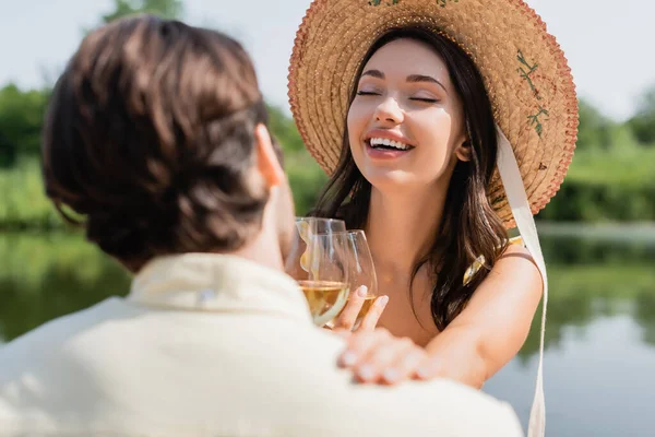 Щаслива жінка в солом'яному капелюсі спирається на розмитого хлопця, тримаючи келих вина — стокове фото