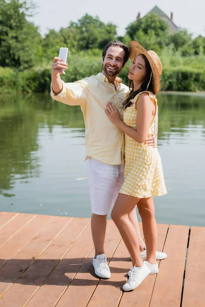 Longitud completa de feliz joven pareja tomando selfie cerca del lago - foto de stock