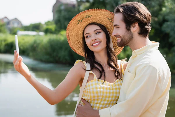 Heureuse jeune femme en chapeau de paille prendre selfie avec petit ami près du lac — Photo de stock