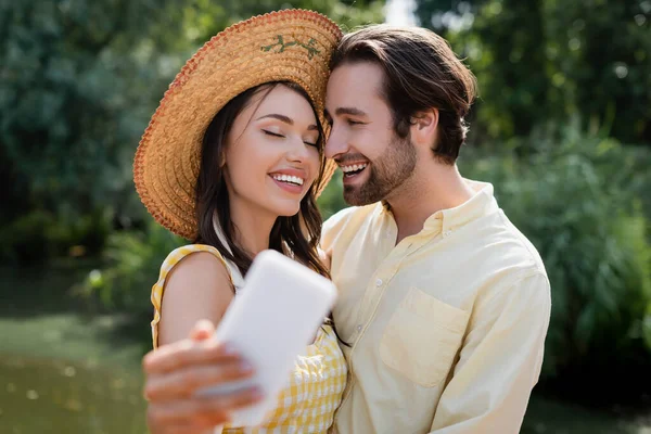 Feliz jovem mulher em chapéu de palha tomando selfie com namorado alegre ao ar livre — Fotografia de Stock