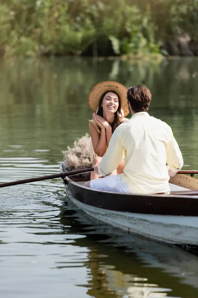 Mujer feliz en sombrero de paja mirando novio romántico durante el paseo en barco - foto de stock