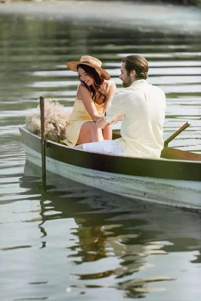 Femme gaie en chapeau de paille tenant la main avec petit ami romantique pendant la promenade en bateau — Photo de stock