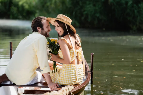 Feliz joven mujer en sombrero de paja celebración ramo de flores cerca de novio alegre durante viaje de jabalí - foto de stock