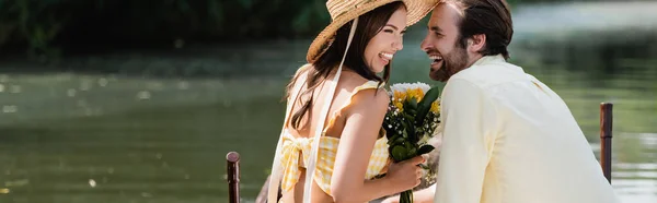 Happy young woman in straw hat holding bouquet of flowers near cheerful boyfriend on lake, banner — Stock Photo