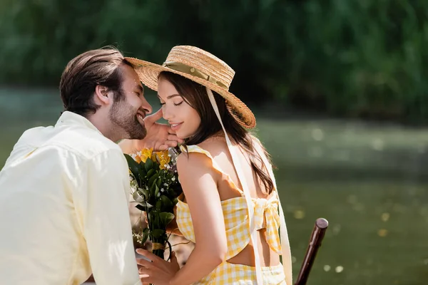 Felice giovane donna in cappello di paglia che tiene bouquet di fiori vicino fidanzato romantico sul lago — Foto stock