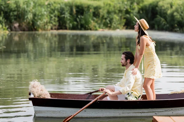 Vue latérale de jeune femme heureuse en chapeau de paille debout derrière l'homme lors d'un voyage romantique en bateau — Photo de stock