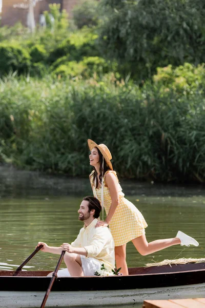 Joyeuse jeune femme en chapeau de paille debout derrière l'homme lors d'un voyage romantique en bateau — Photo de stock