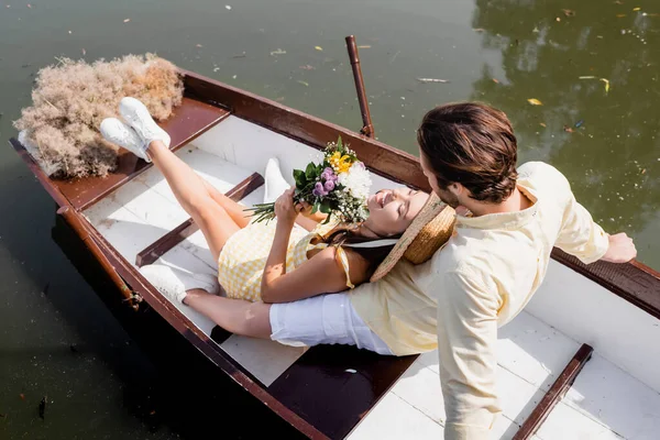 High angle view of young woman in straw hat holding bouquet of flowers and lying in boat with man — Stock Photo