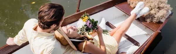 Visão de alto ângulo da jovem mulher em chapéu de palha segurando buquê de flores e deitado em barco com o homem, bandeira — Fotografia de Stock