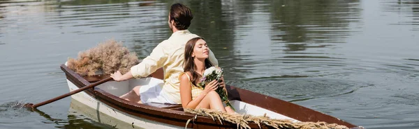 Молода жінка тримає квіти і спирається на хлопця під час романтичної подорожі на човні, банер — стокове фото