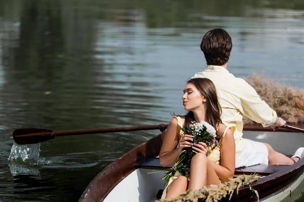 Young woman holding flowers and leaning on back of man during romantic boat trip — Stock Photo