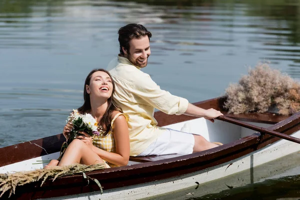 Feliz jovem segurando flores e inclinando-se na parte de trás do namorado durante a viagem de barco romântico — Fotografia de Stock