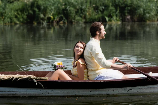 Feliz joven mujer sosteniendo flores y apoyándose en la espalda de novio barbudo durante romántico viaje en barco - foto de stock