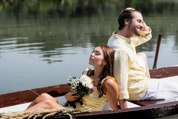 Счастливая молодая женщина с цветами и опираясь на спину улыбающийся парень во время романтической поездки на лодке — стоковое фото