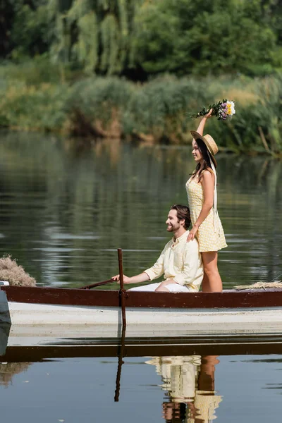 Vue latérale de la femme heureuse tenant des fleurs et debout derrière l'homme dans le bateau — Photo de stock