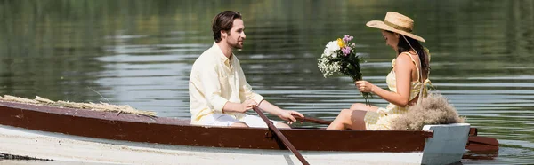 Vue latérale de la jeune femme en chapeau de paille tenant des fleurs et ayant romantique promenade en bateau avec l'homme, bannière — Photo de stock