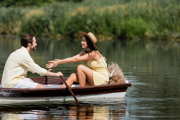 Smiling woman in straw hat sitting out outstretched hands near man in boat — Stock Photo